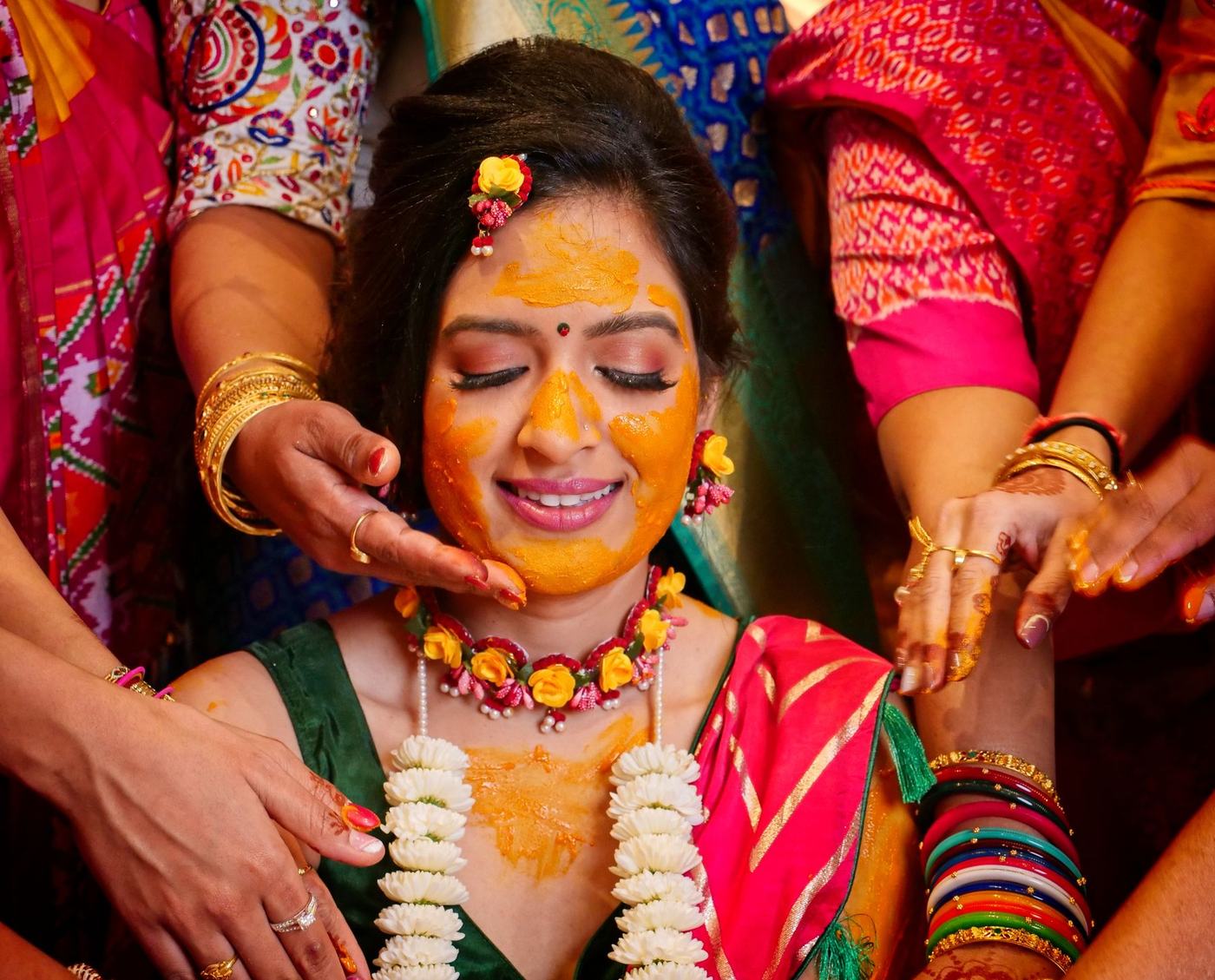 Bride with eyes closed as family hands apply haldi