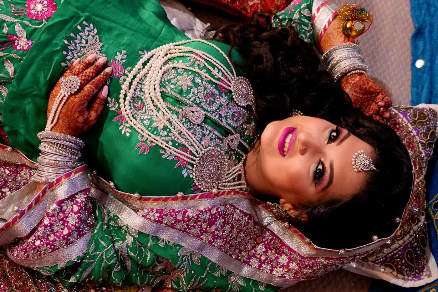 Bride in an emerald lehenga lying back among her jewels and mehndi