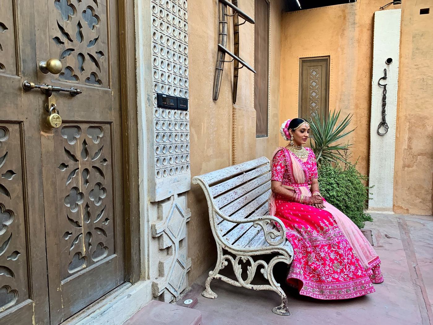 Bride in a pink lehenga on a bench beside carved wooden doors