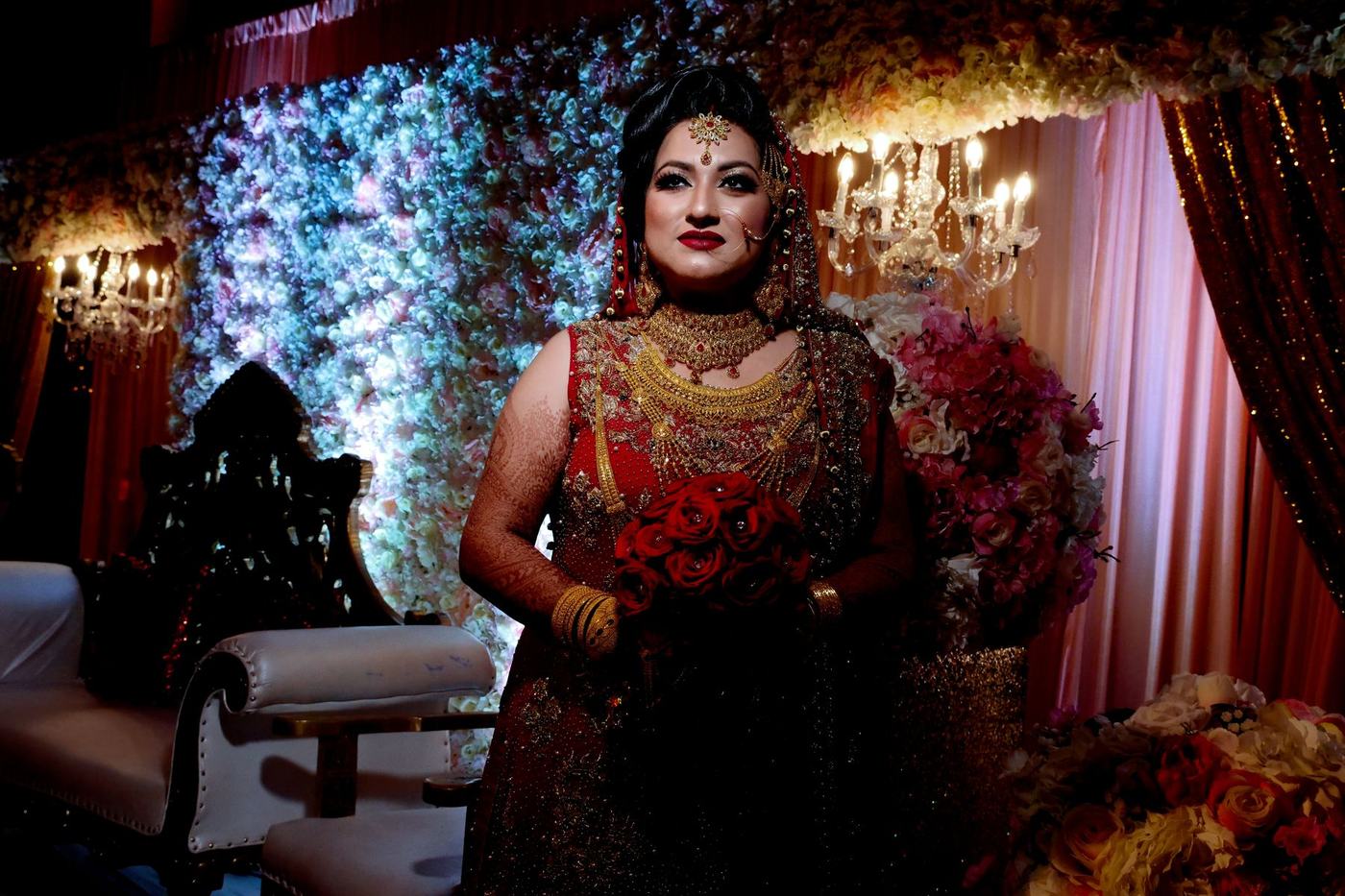 Bride in red before a wall of reception flowers and chandeliers