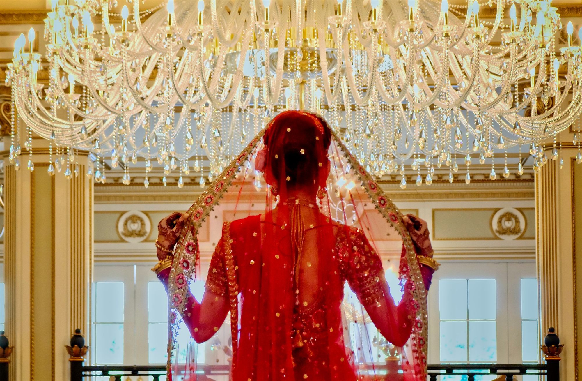 Bride lifting her red veil beneath a ballroom chandelier