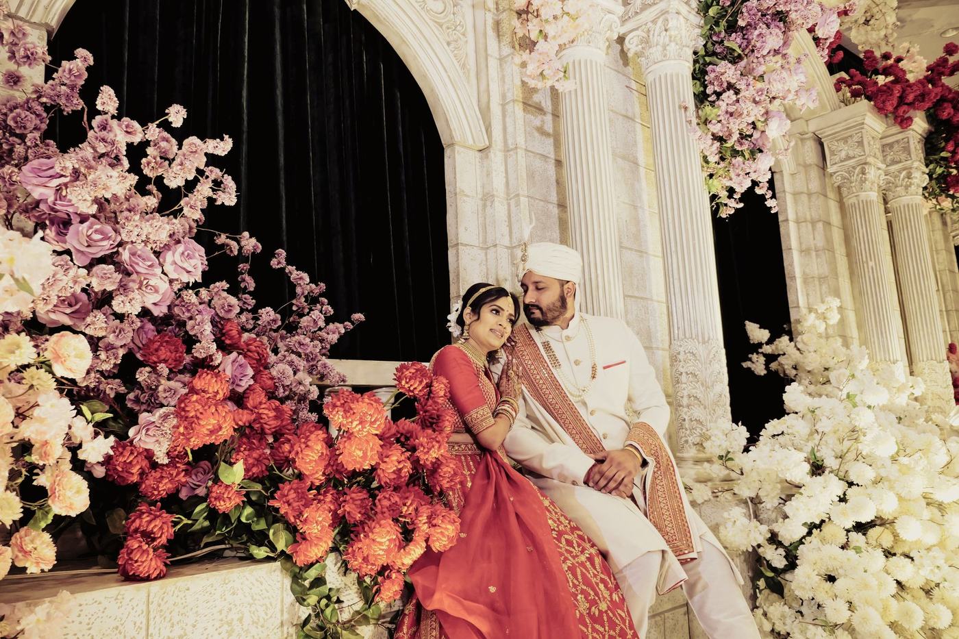 Couple seated among blush and crimson florals on the reception stage