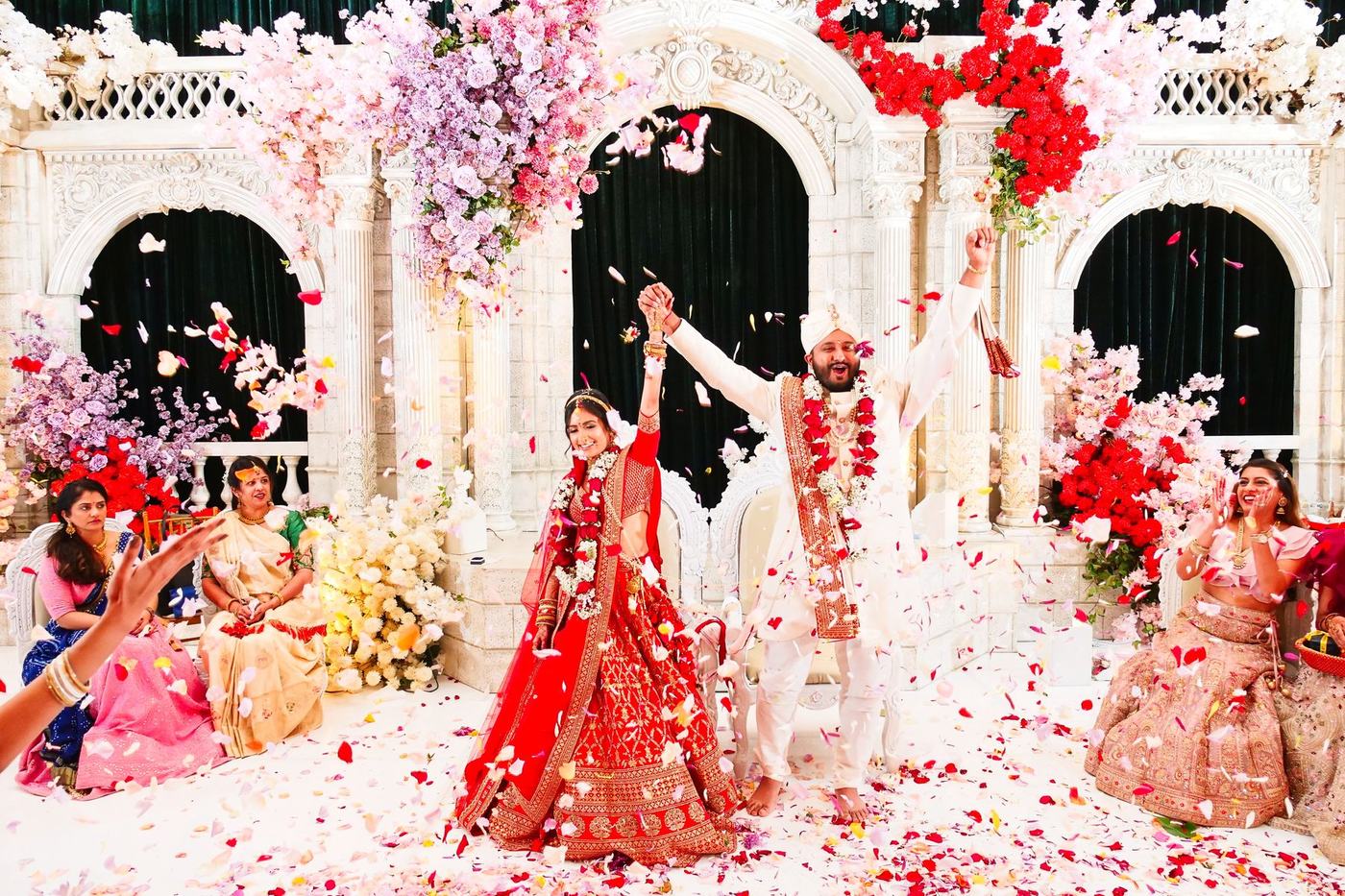 Couple laughing in a storm of petals at the white mandap